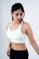 A person in a white sports bra and black leggings stands against a plain background, looking down. The sports bra has the text "Push Your Limits.